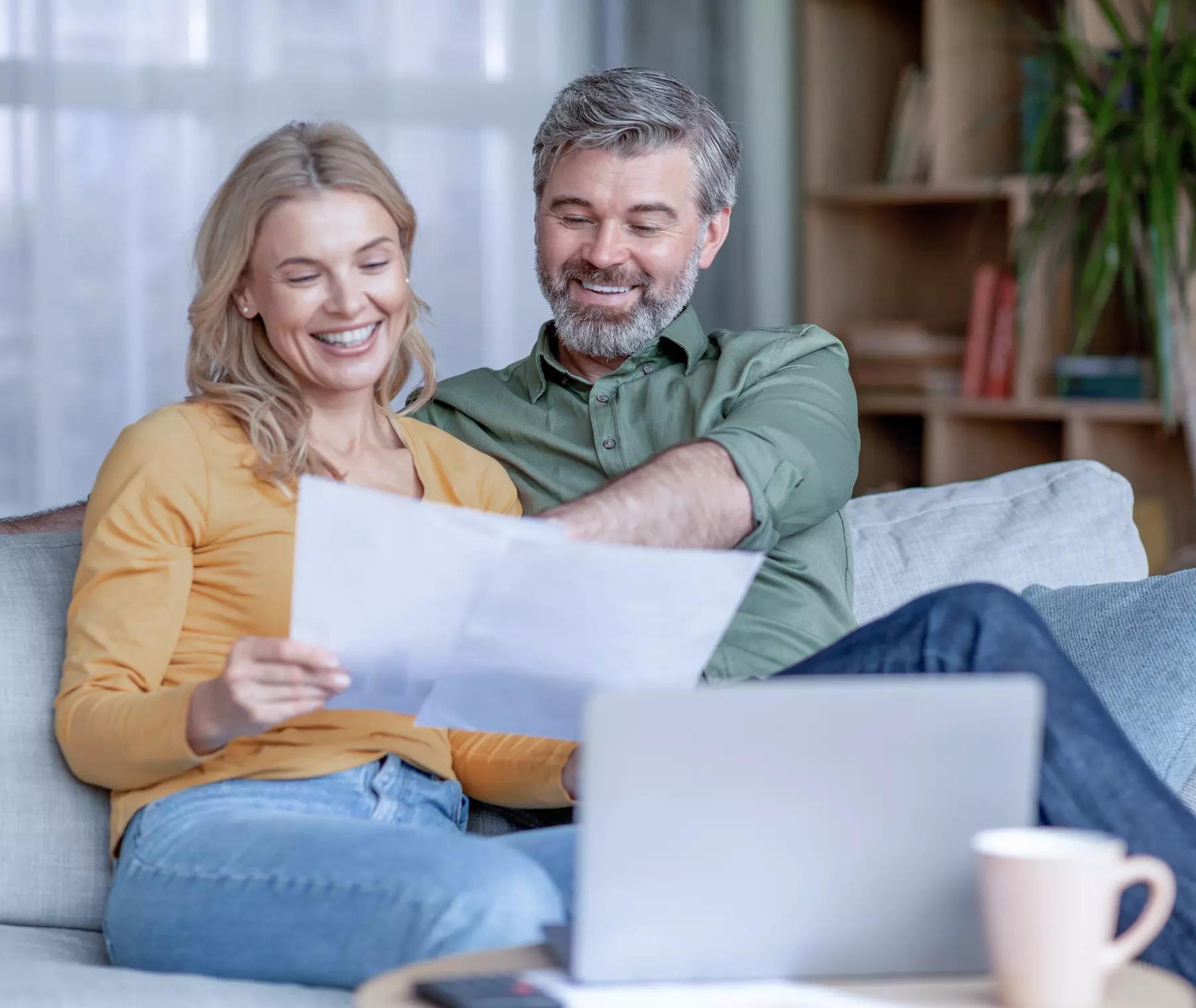 Happy Middle Aged Couple Checking Financial Papers Downloaded from the Generosity Calculator While Sitting Together On Couch In Living Room