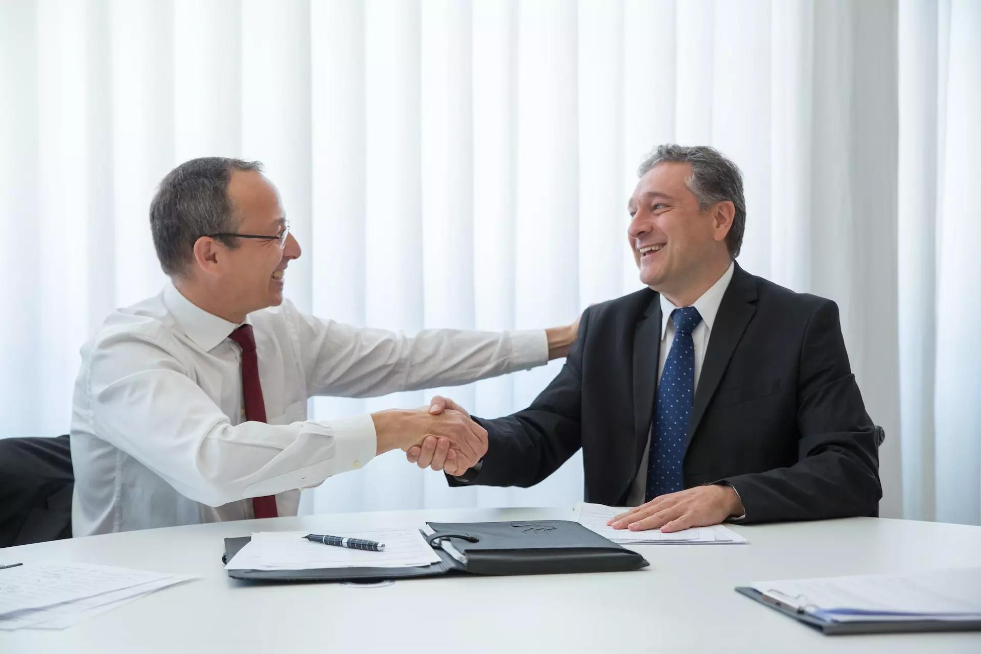 two men at a table in an office setting shake hands